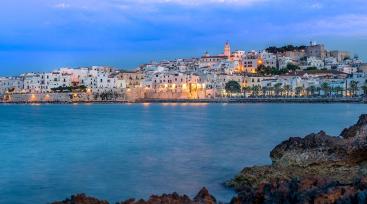 Vista serale di un borgo costiero illuminato, con mare calmo e cielo azzurro.